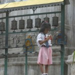 A little girl stands next to some prayer wheels in a Kathmandu courtyard